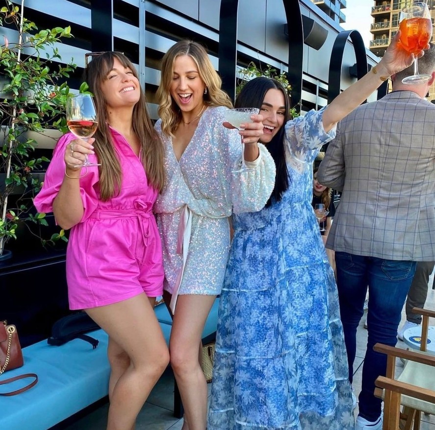 Three friends in a pink playsuit, a sequin wrap dress and a blue floral maxi raising their glasses on a rooftop
