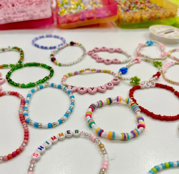 Letter bead friendship bracelets spread across a table, spelling TAYLOR, RED and SHIMMER among rainbow and seed bead strands