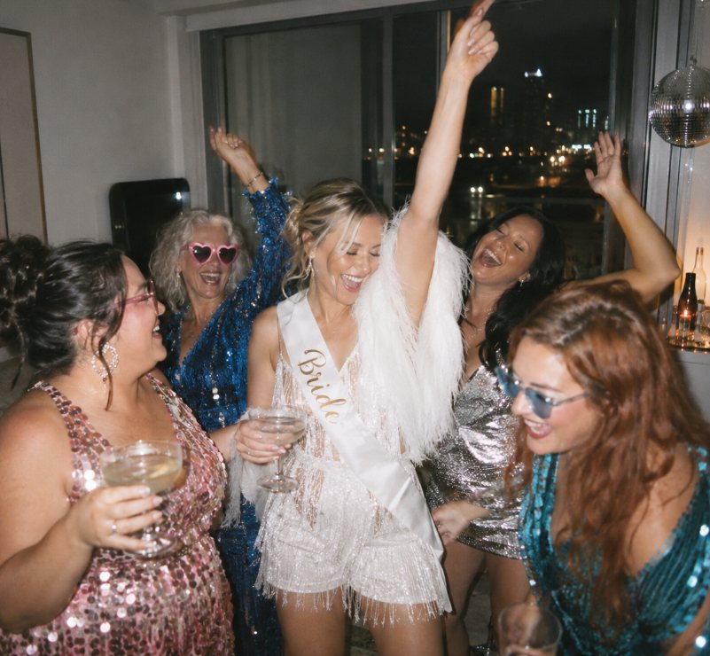 A bride in white fringe and feathers dancing with her arm up, her friends in sequins dancing around her