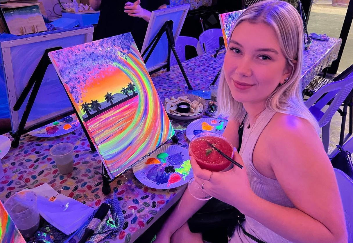 A guest smiling over her shoulder with a frozen strawberry cocktail beside her glowing rainbow wave painting
