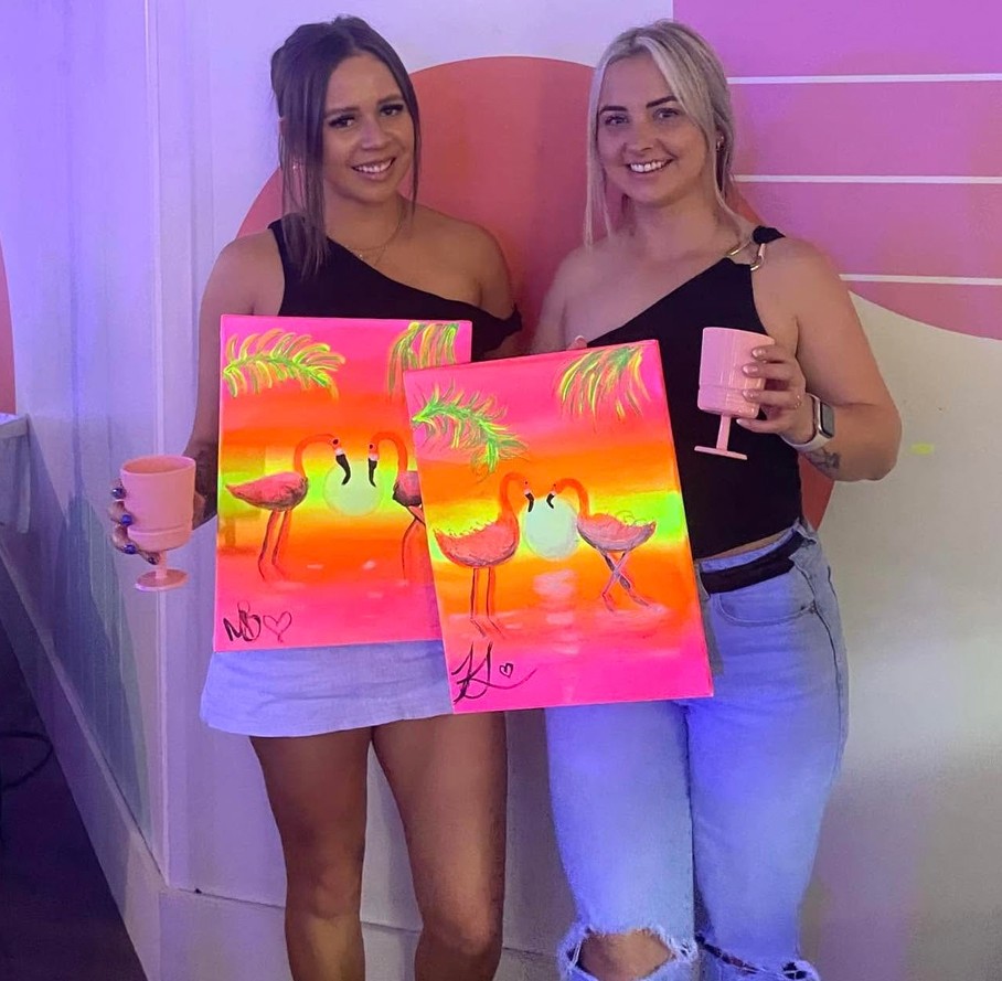 Two friends holding up glowing neon canvases of two flamingos in love