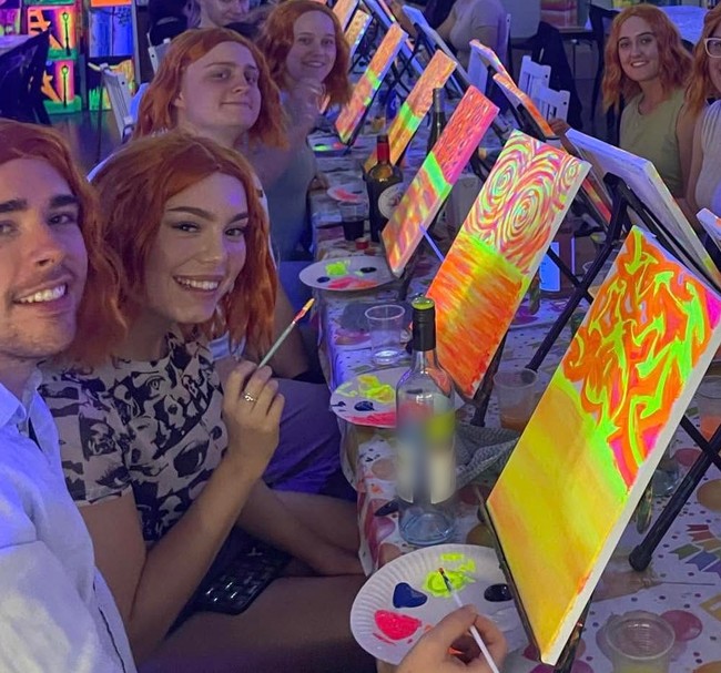 Friends in matching red wigs grinning down a table of glowing neon canvases
