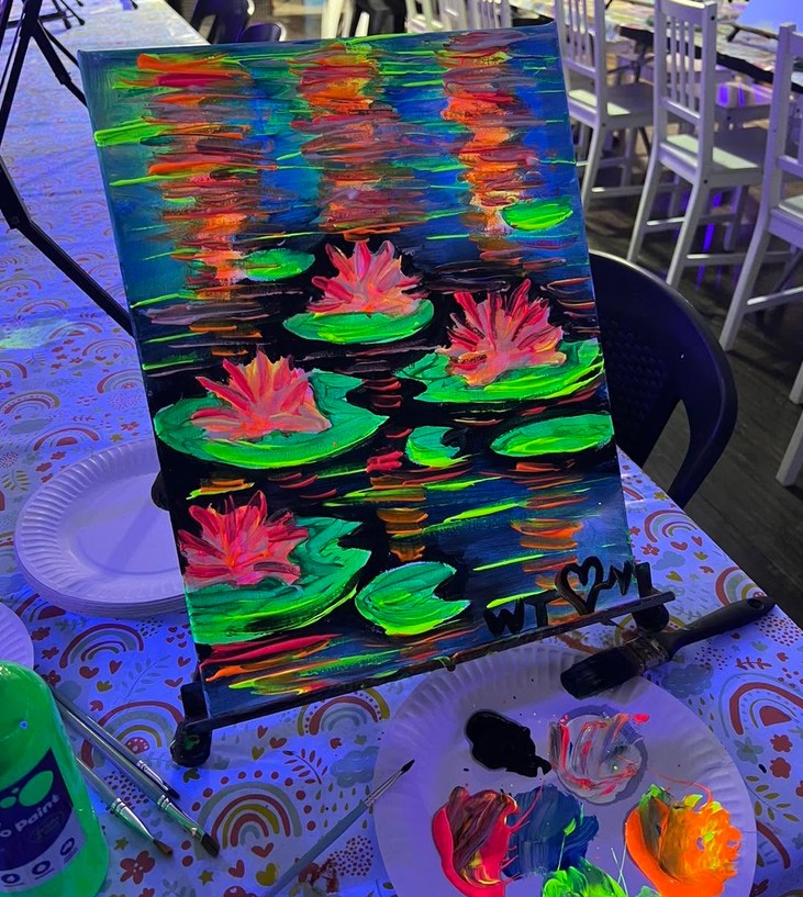 A finished glow painting of water lilies on an easel, bright pink and green under UV light