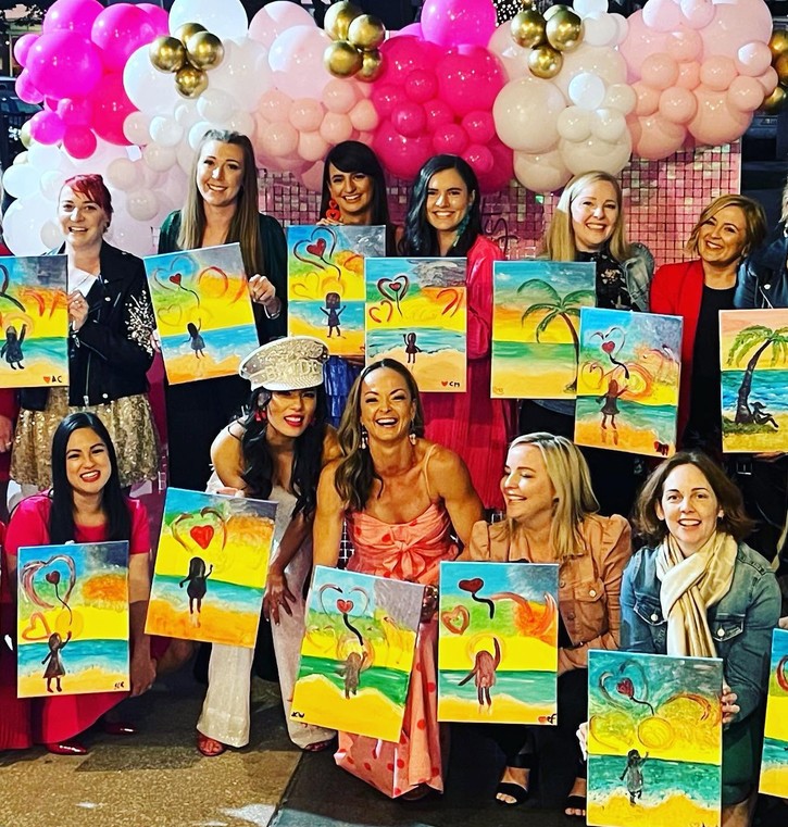 A hens crew under a pink and gold balloon arch holding up their heart paintings, the bride in a Bride cap in the middle