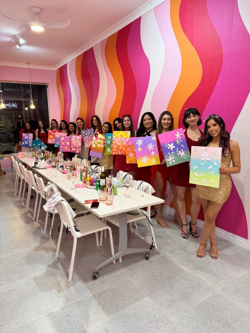 The bride in gold sequins holding her canvas up front, her friends holding theirs behind her along the table