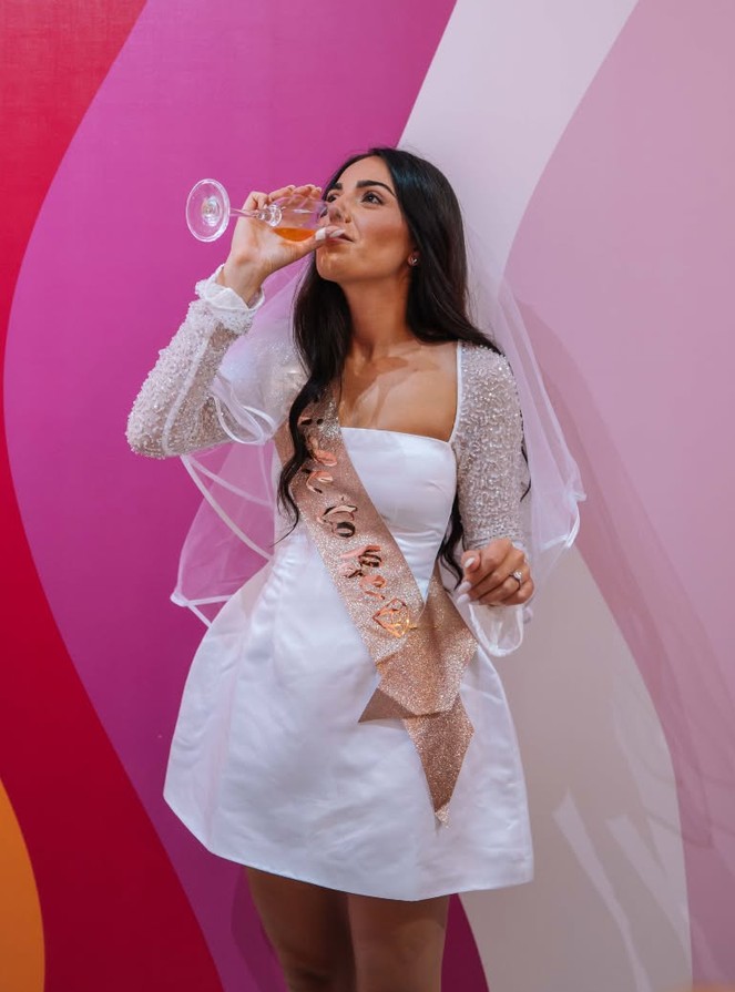 The bride in her veil and sash sipping a drink against the studio's painted pink wall
