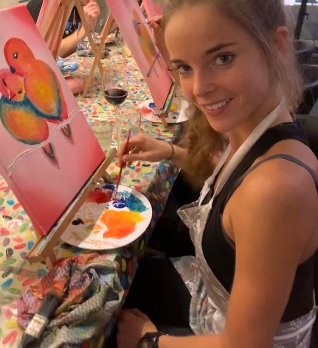 A guest in an apron smiling beside her painting of two lovebirds, a glass of red at her elbow