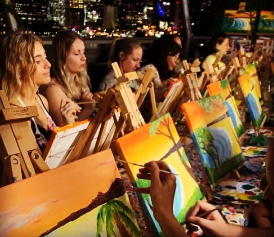 A row of guests at easels painting bright sunsets, the city lights glowing behind them at night