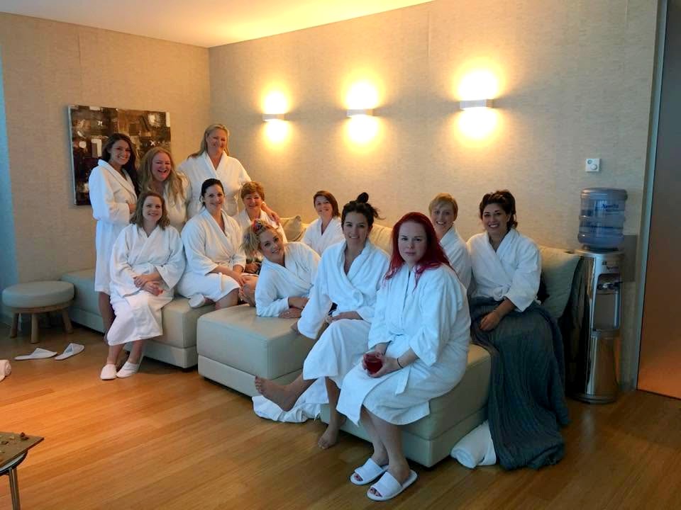 A big hens crew of thirteen in white robes and slippers on the sofas of a warm spa lounge