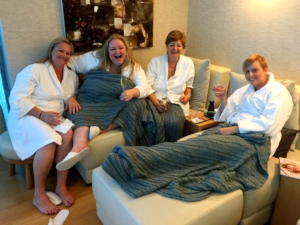 Four friends in robes curled up under knitted grey blankets on a cream lounge, glasses of bubbles in hand, one laughing