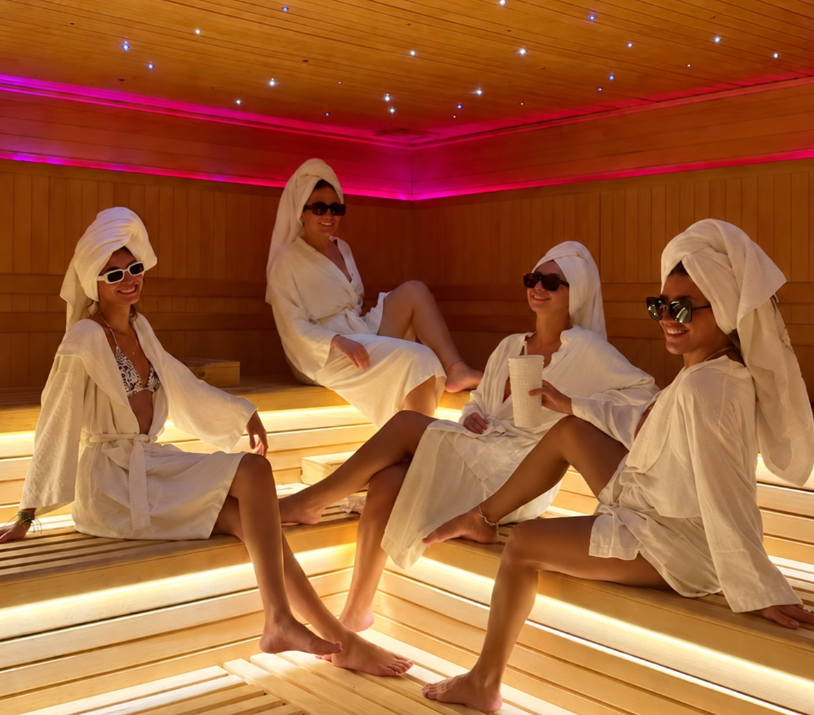 Four hens in robes and towel turbans lounging on the benches of a sauna lit pink, little star lights in the ceiling