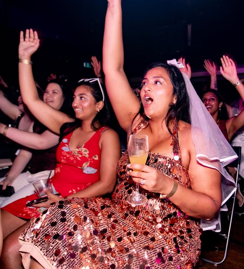 The bride in her veil and a sequinned dress cheering with her arm in the air, the whole crowd behind her doing the same