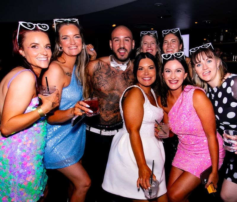 A hens crew in heart sunglasses around the bride in white and a tattooed performer in a bow tie