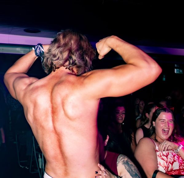 A performer flexing with his back to the camera while the hens in the front row scream with laughter