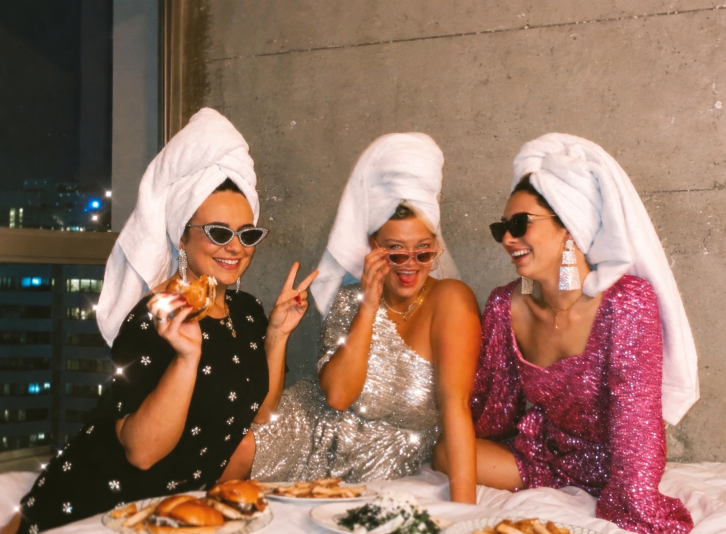 Three friends in towel turbans and sequins laughing over room service in a hotel room