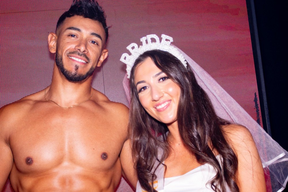 The bride in a BRIDE tiara and veil smiling beside her topless waiter