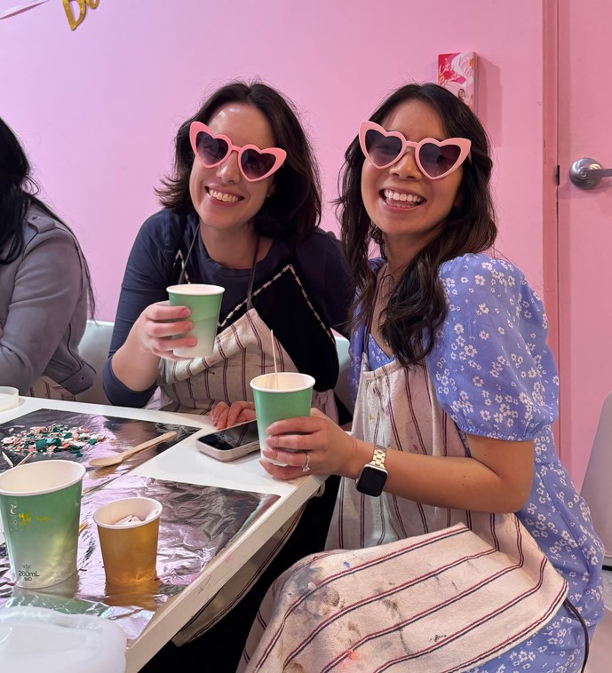 Two friends in aprons and pink heart sunglasses grin over their cups at a craft table