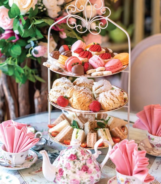 A three-tier stand of sandwiches, scones and macarons beside a floral teapot and pink napkins in floral teacups