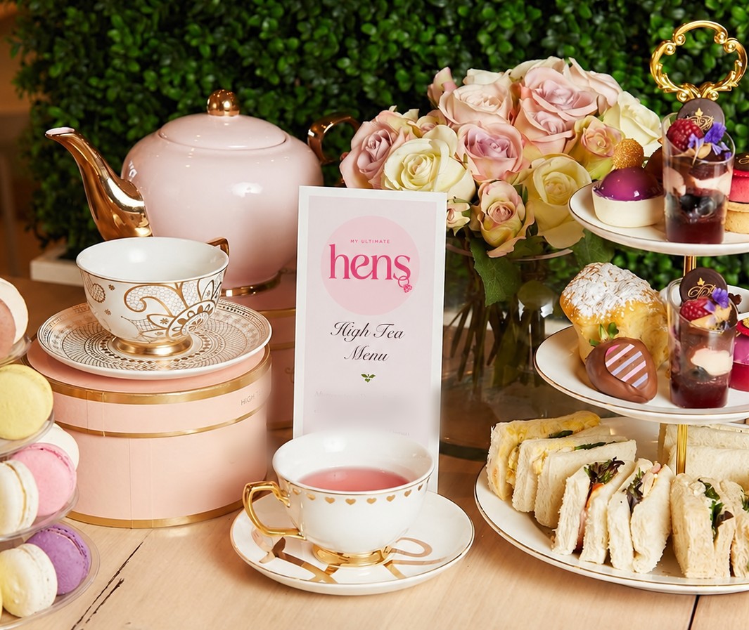 A pink teapot, gold-rimmed teacups, roses and a My Ultimate Hens high tea menu beside a tiered stand of sandwiches and sweets