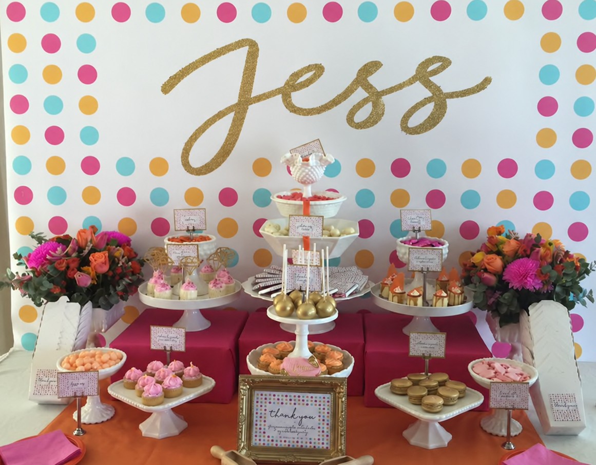 A dessert table of tiered stands, gold macarons and cakes in front of a polka dot wall with the bride's name in gold glitter