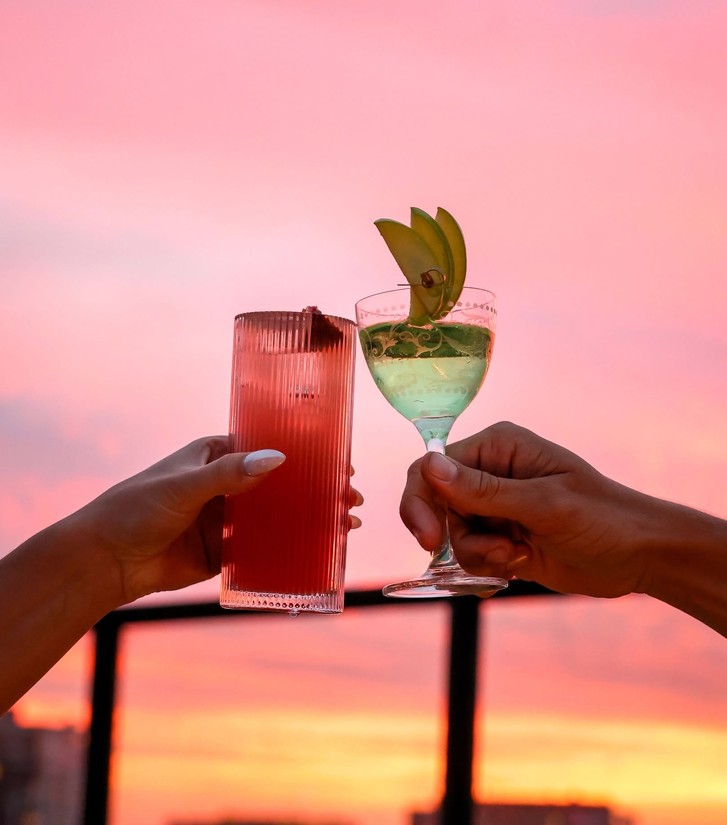 Two cocktails clinking against a pink sunset sky on a rooftop