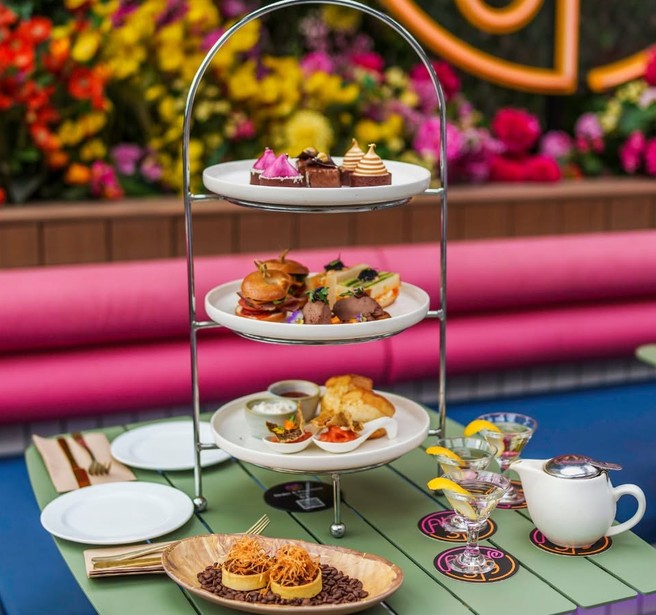 A high tea table set with a three-tier stand, a teapot and cocktails in front of a flower wall