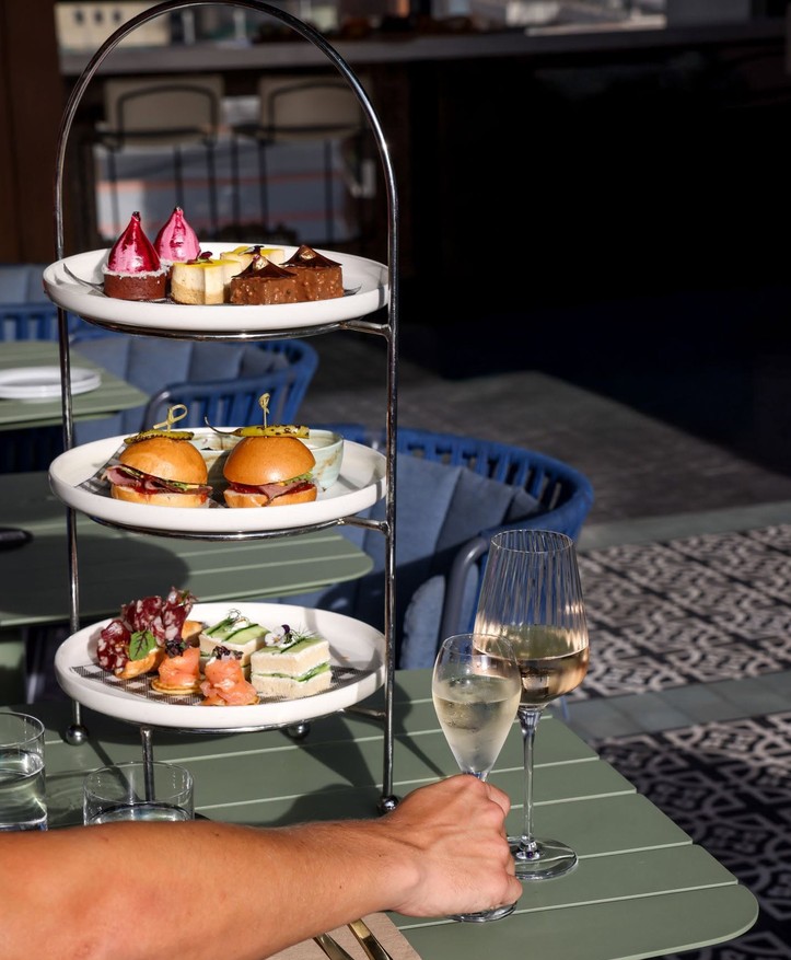 A three-tier high tea stand of savoury bites, sliders and little cakes, with glasses of bubbly on the table