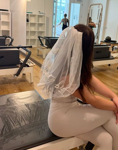 The bride in a short veil and a pale bodysuit sitting on a reformer in a pilates studio
