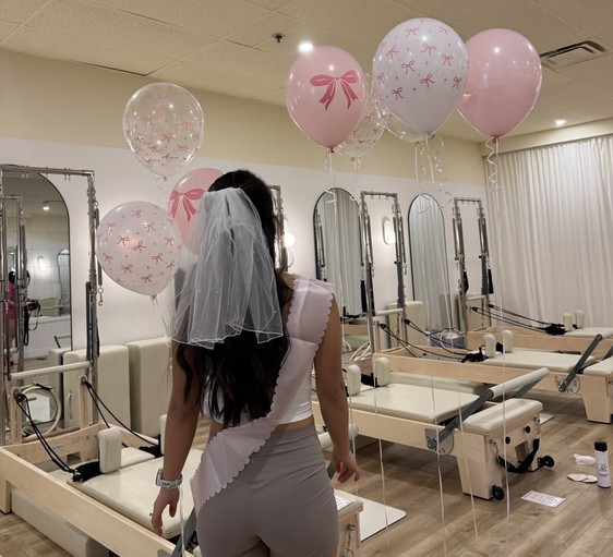 The bride in a veil and sash looking across a reformer studio decorated with pink bow balloons