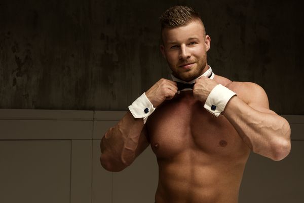 A shirtless waiter in a bow tie and cuffs smiling at the camera