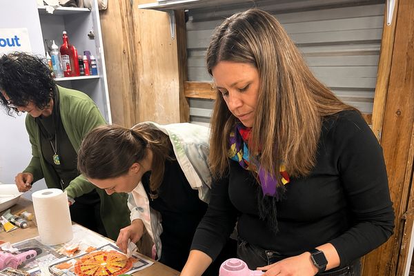 Three women finishing their mosaic pieces at a workshop table with drinks nearby
