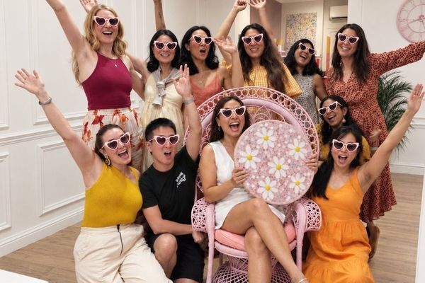 Bride in a pink peacock chair holding a pink and white daisy mosaic, with her friends in heart sunglasses cheering around her