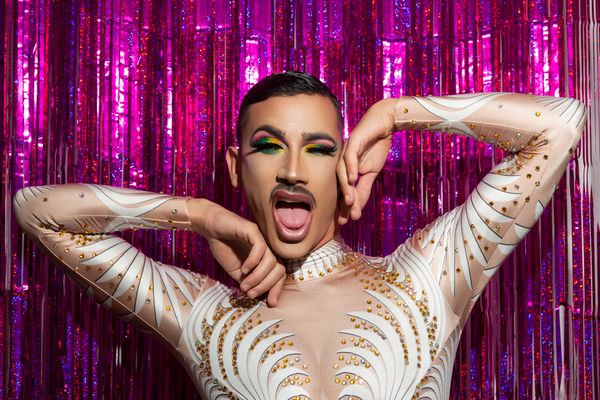 A drag performer in a jewelled bodysuit pulling a face in front of a pink tinsel curtain