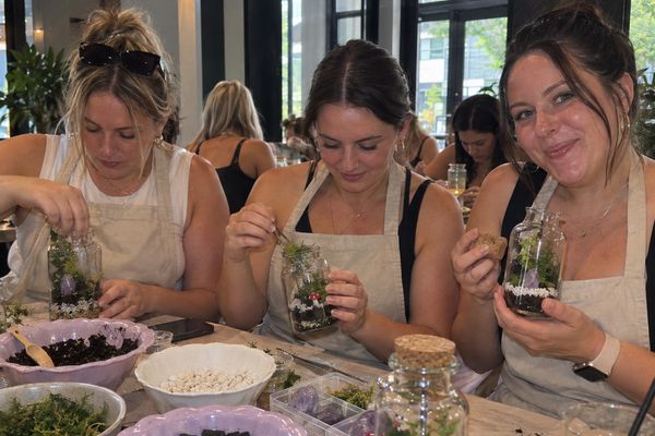 Three friends in aprons plant tiny gardens in glass jars at a workshop table