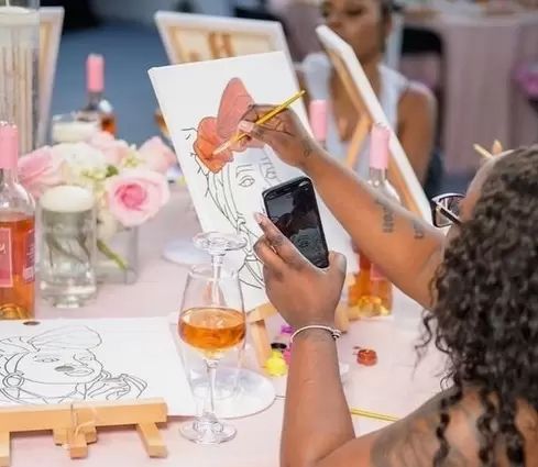 A hen photographing her portrait canvas at a paint and sip table with roses and wine
