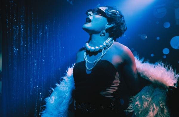 A drag queen in pearls and a feather boa singing under blue stage lights