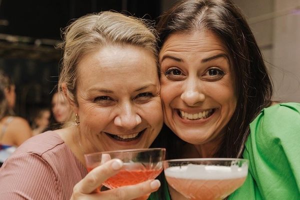 Two friends cheek to cheek, raising pink cocktails in coupe glasses
