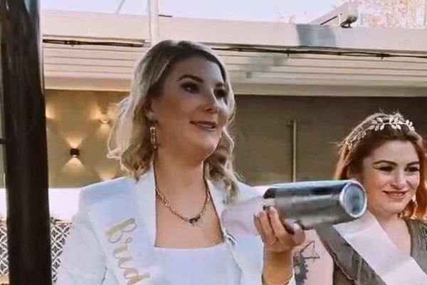 Bride in a white sash shakes a cocktail beside a smiling friend wearing a tiara