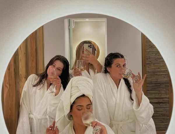 Friends in white robes and a towel turban sipping bubbly in front of a round mirror