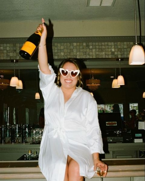 A woman in a white shirt dress and heart sunglasses holding a bottle of bubbles over her head