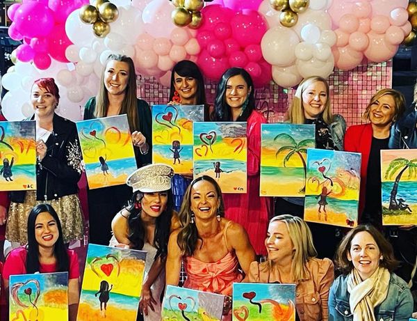 The bride and her crew holding up their beach paintings with hearts in front of pink and gold balloons