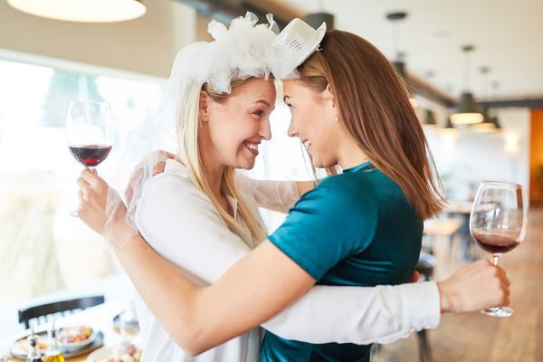 A bride to be in a feather headpiece touching foreheads with a friend over glasses of red wine
