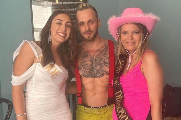 A shirtless fireman host in braces with the bride and a friend in a pink hat