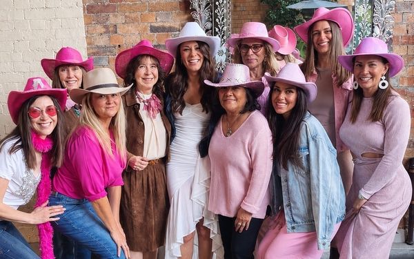 Hens of all ages in pink cowgirl hats smiling together in a brick courtyard