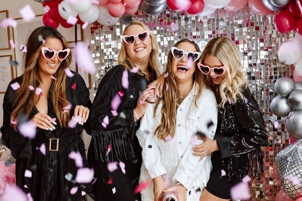 Four friends in heart sunglasses laughing under confetti, the bride in white fringe