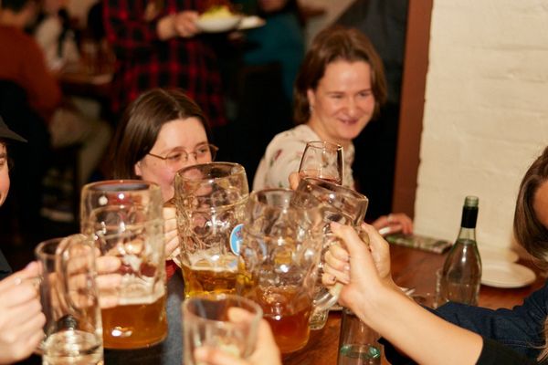 Friends clinking big mugs of beer across a busy beer hall table