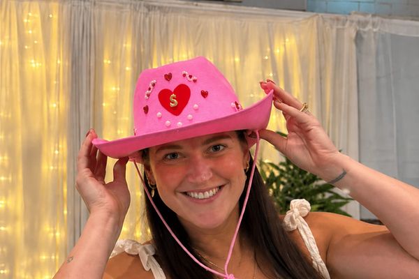 A smiling woman in an apron shows off the pink cowboy hat she painted