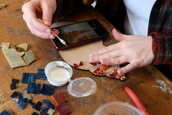 Hands pressing small glass tiles onto a mosaic base beside a phone showing the design