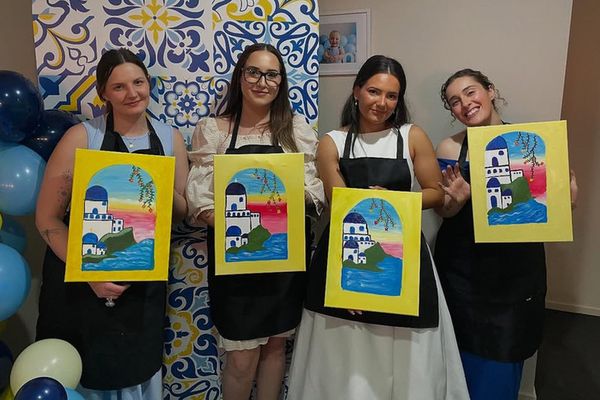 Four friends in aprons hold up their painted canvases after a paint class