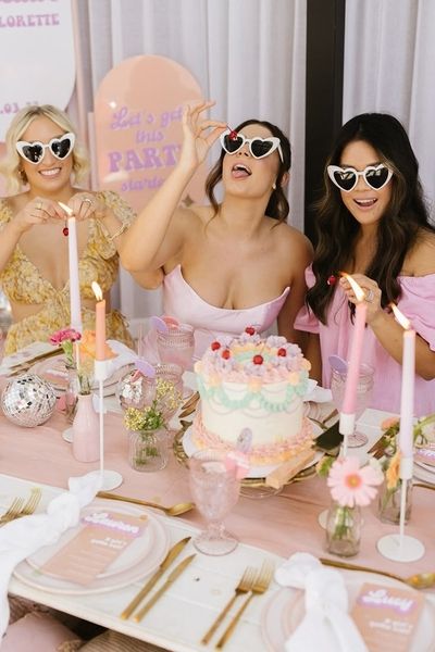 Three friends in heart sunglasses at a pink table with a cake and candles